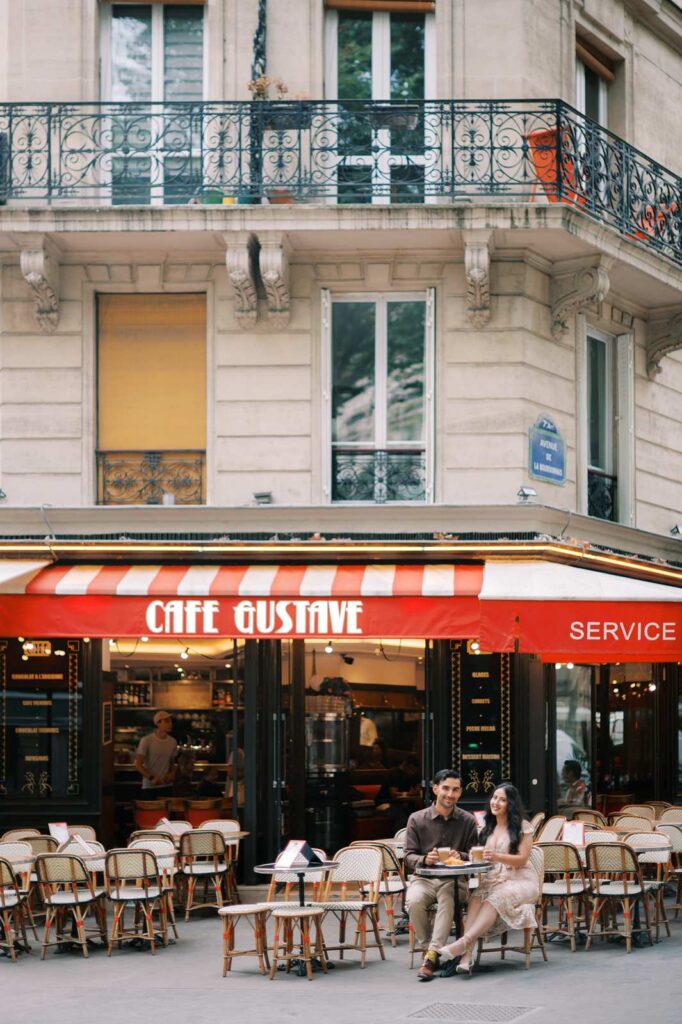 A couple sits at an outdoor table under a red and white striped awning at Café Gustave, celebrating their 4th wedding anniversary in the 7th arrondissement of Paris. They are smiling and enjoying drinks, surrounded by other empty tables and classic wicker café chairs.