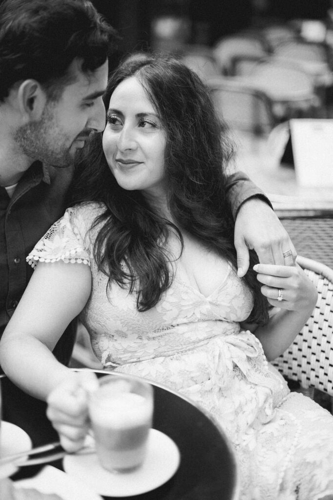 A black-and-white photo of a couple sitting closely together at a table in an outdoor café in Paris's 7th arrondissement. One person holds a latte in one hand, and her husband's hand in the other. She looks up at him affectionately, and he smiles down at her with his arm around her shoulder.