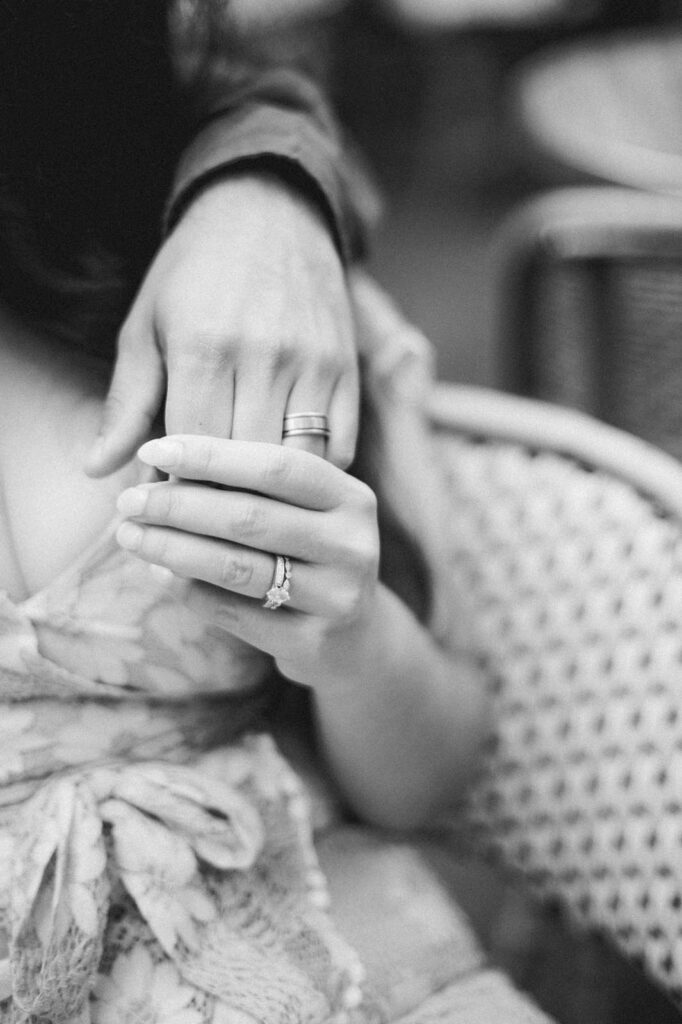 Black and white close-up of a married couple during an anniversary photo session. One person's left hand is around their partner's left shoulder, and as they hold hands we see both of their wedding rings. The focus is on their intertwined hands and wedding rings, and the background is blurred.