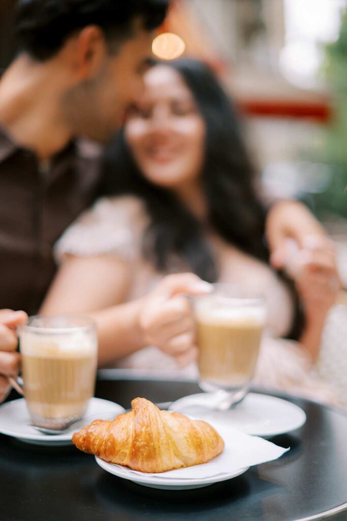 A couple cuddles at a Paris café, each holding a latte. The couple themselves are blurred, and a croissant on a napkin and plate is in focus in the foreground.
