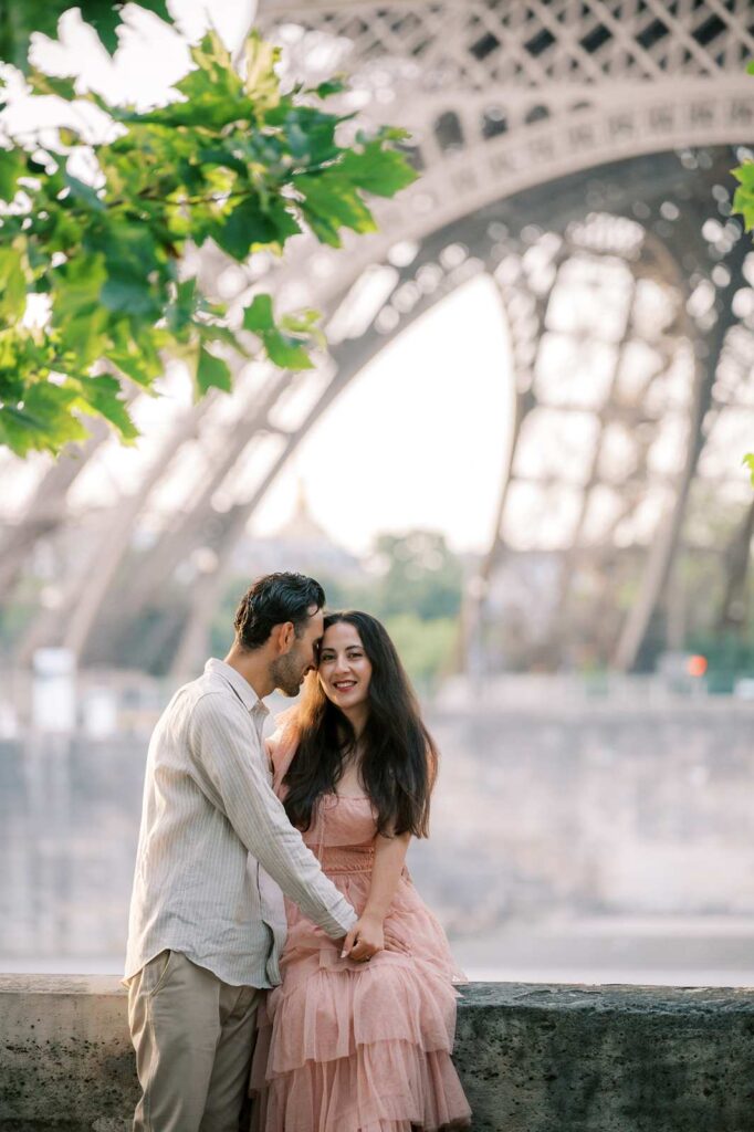 A husband gets close and cozy with his wife as she smiles at the camera while sitting on a ledge in front of the Eiffel Tower. Sunlight filters through green leaves overhead, adding a romantic ambiance to their 5th anniversary photo shoot.