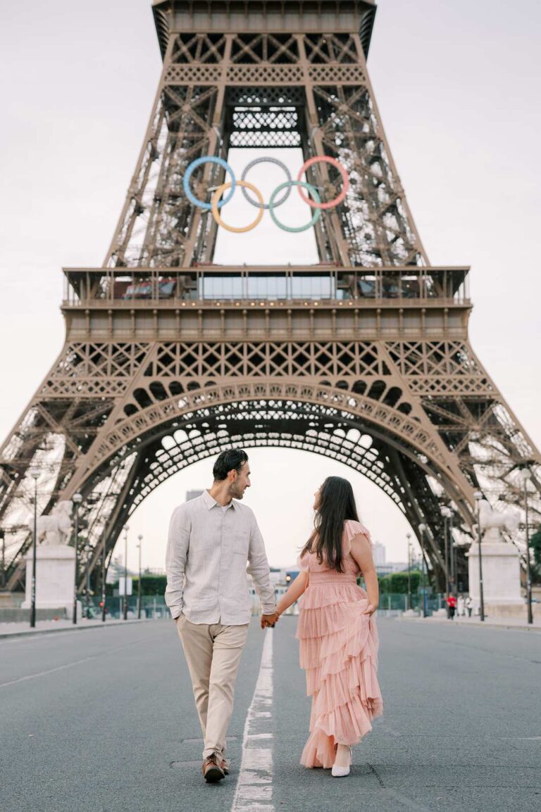 A couple holding hands and walking away from the Eiffel Tower during their romantic 5th anniversary photo session. They are looking back over their shoulders at the Olympic rings on the Tower for the 2024 Olympics. The man is dressed in an offwhite shirt and khaki pants, and the woman is wearing a flowing pink dress.