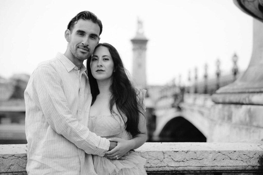 A black-and-white photo of a couple hugging each other and smizing on the Pont Alexandre III bridge in Paris. The are both looking straight at the camera for a serious and slightly posed photo during their 5th anniversary photo shoot.