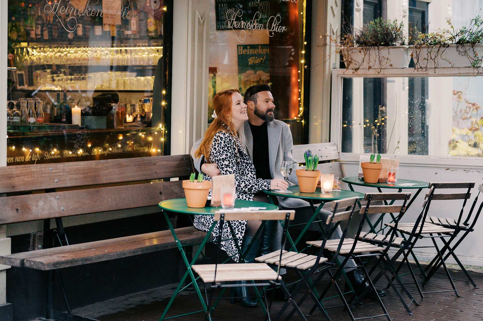 A couple sits at an outdoor café table, holding hands, surrounded by lit string lights and potted plants. This intimate Jordaan photo session captures their romance in the neighborhood's charming ambiance.