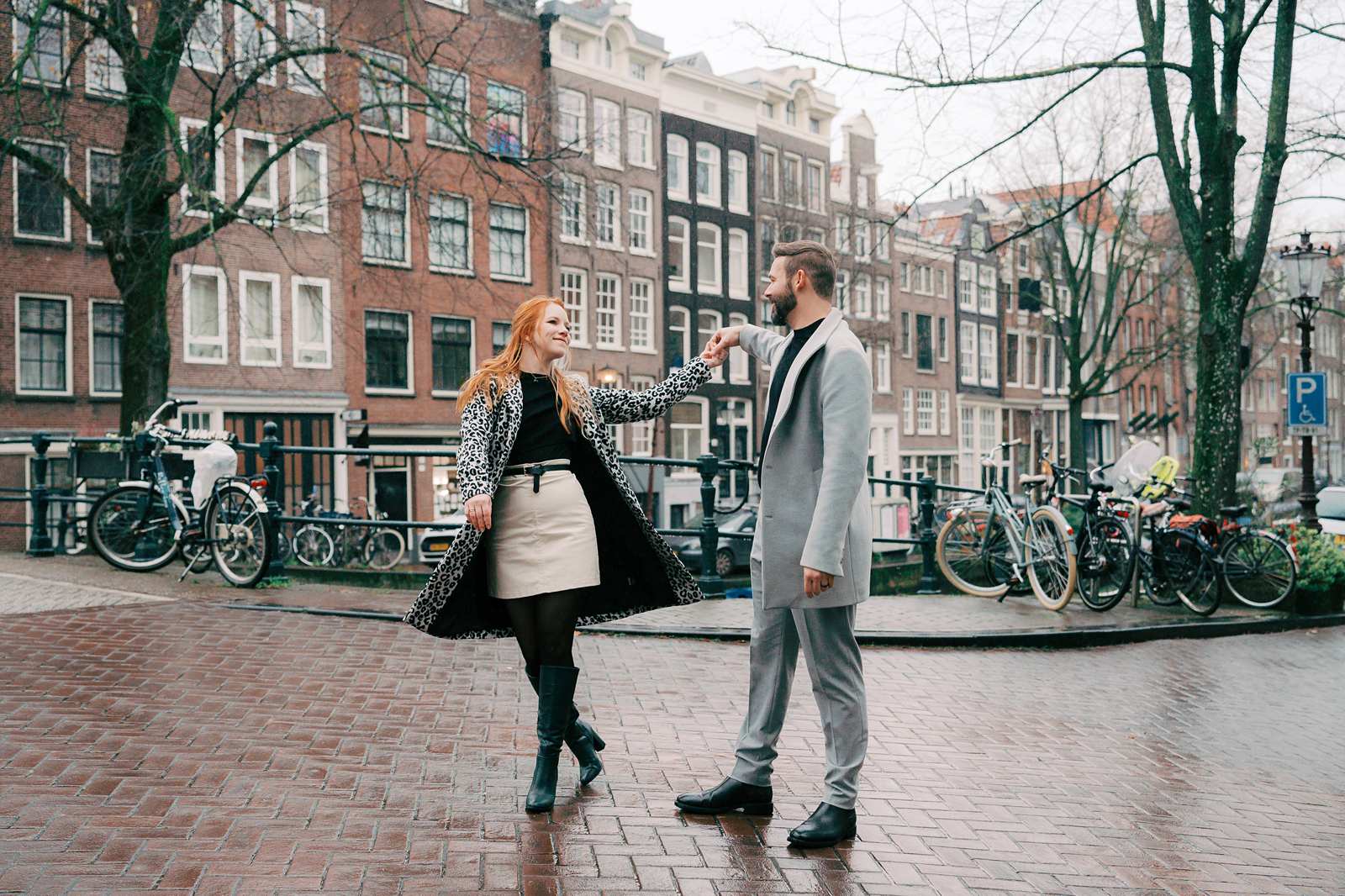 A couple dances in the middle of a wet brick street in the Jordaan neighborhood of Amsterdam during their Amsterdam photoshoot, with rows of bicycles and traditional narrow canal houses behind them.