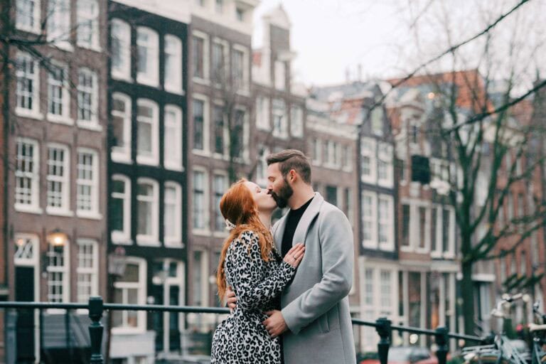 A couple kissing on a canal bridge in the Jordaan neighborhood of Amsterdam, with tall, narrow canal houses blurred in the background.