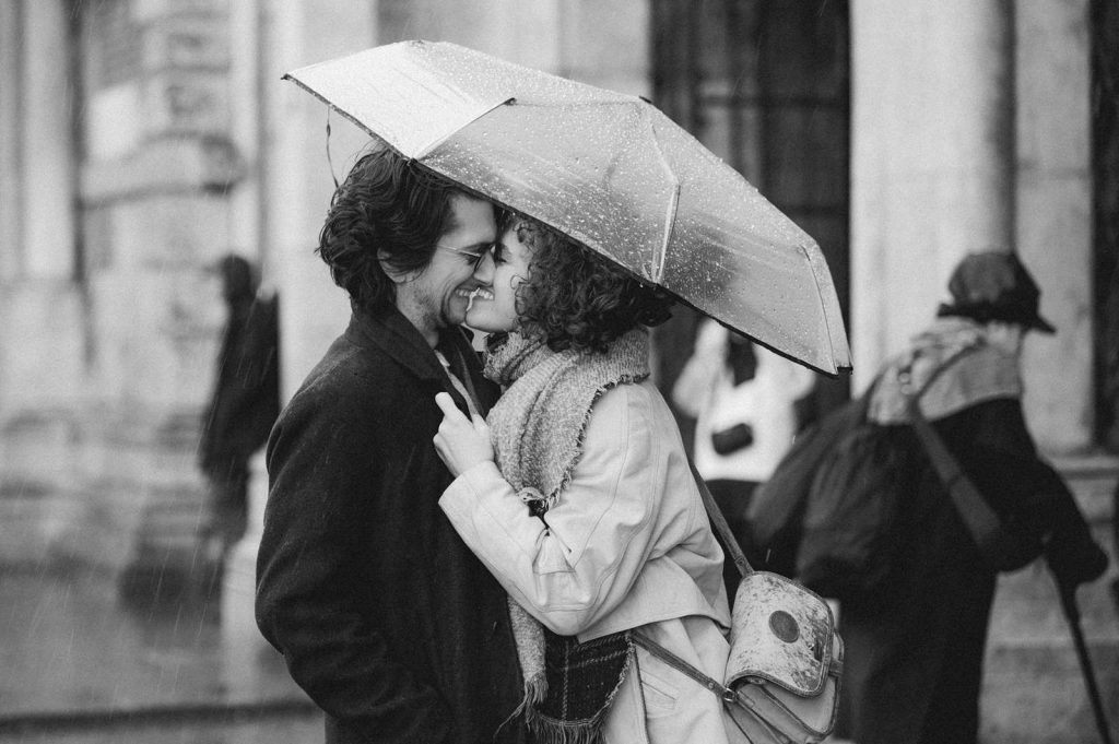 A person pulls her partner to her by his lapels while they share an umbrella in the rain. With lips almost touching, they smile as blurred people walk past in the background.