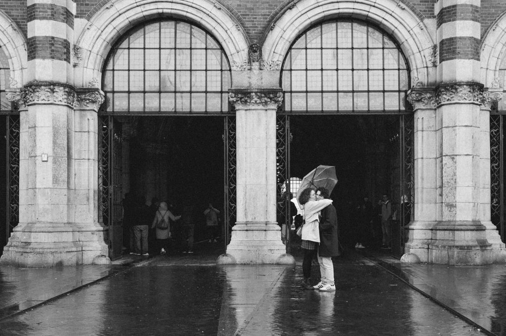 Black and white photo of a couple under a shared umbrella umbrella. On person stands on tiptoes with arms around the other's shoulders, giving him a kiss on the cheek. They're in front of the large, arched entrance to the Rijksmuseum on a rainy day, during a romantic photoshoot.