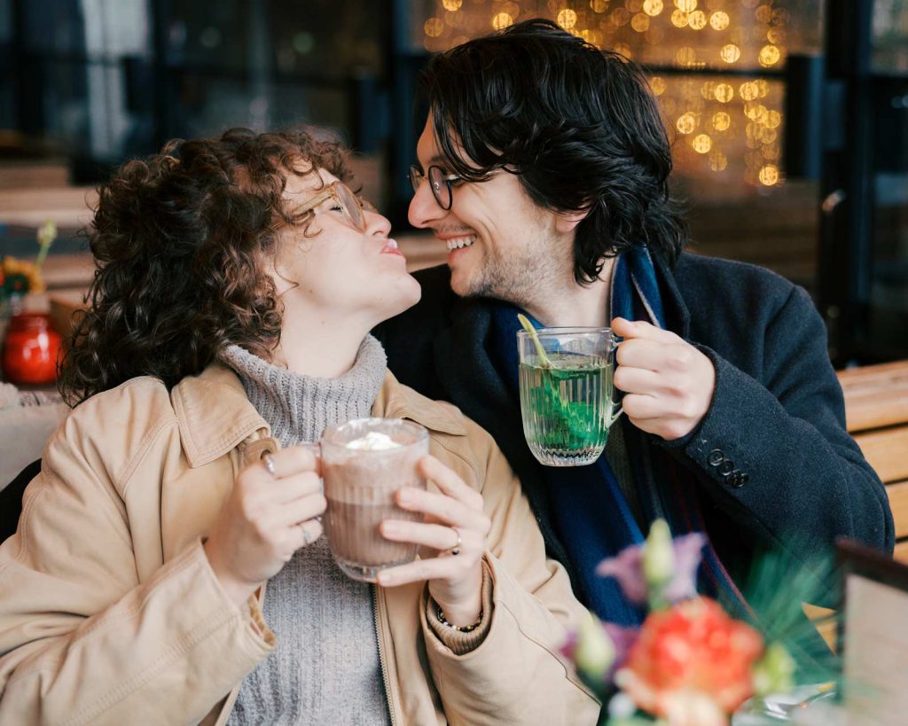 A couple toasts with hot drinks and laughs as one partner moves to kiss the other with milk foam on her upper lip. They're at an outdoor cafe in Amsterdam, surrounded by warm, decorative lights.