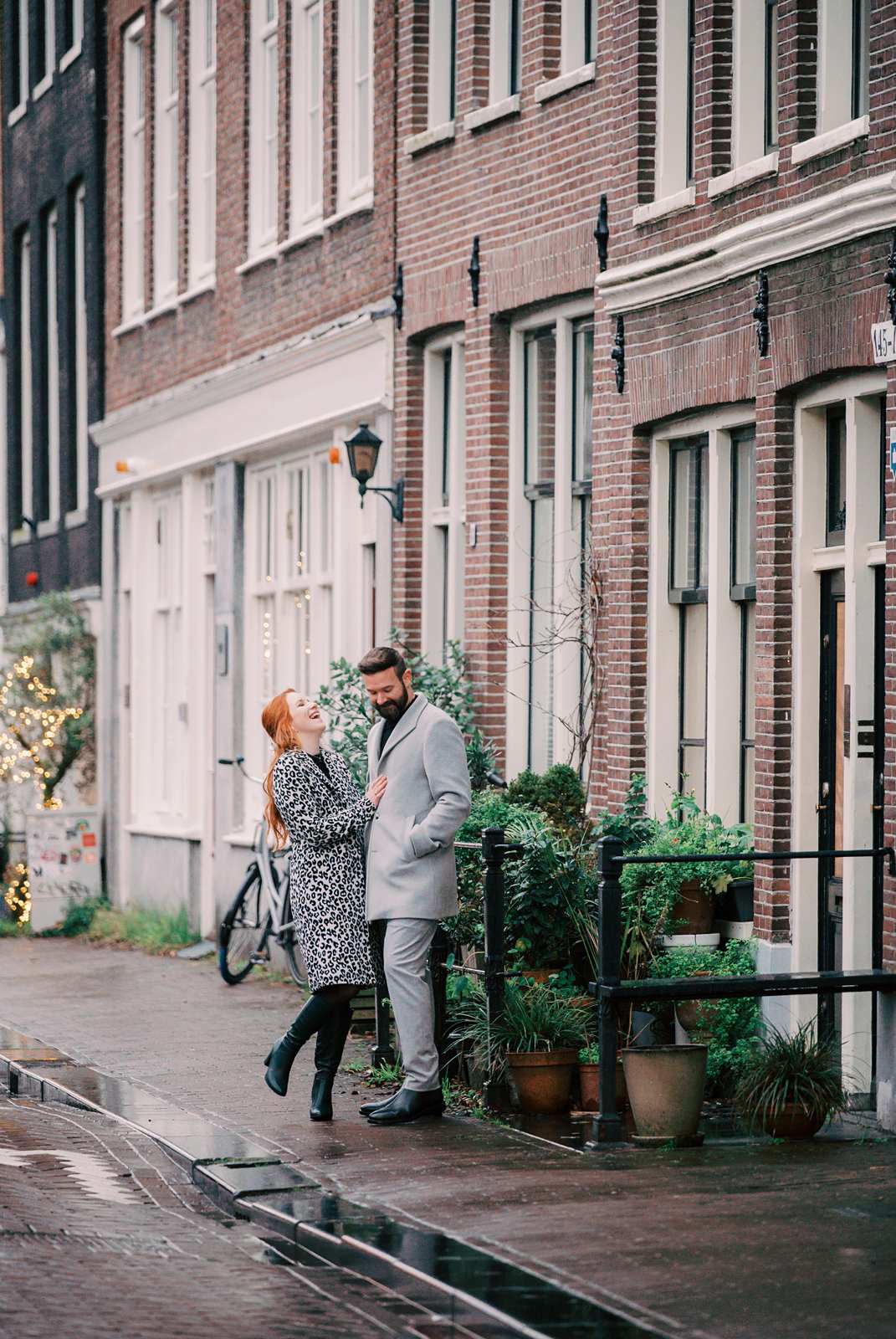 A couple stands laughs together on a rainy street in the Jordaan neighborhood, surrounded by potted plants and brick buildings.