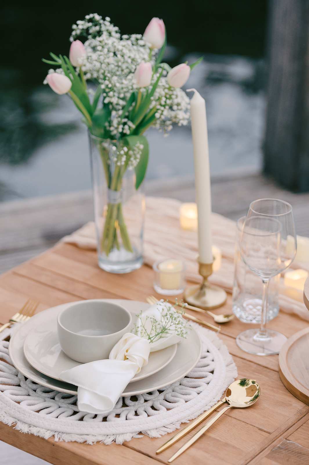 Elegant table setting with white dishes, gold cutlery, flowers, and candles on a wooden table by the water—perfect for a luxury picnic or an Amsterdam proposal photo shoot.