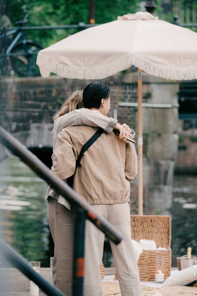Two people in beige outfits hug by a canal in Amsterdam under a white umbrella with picnic items nearby—a perfect moment for a surprise proposal.