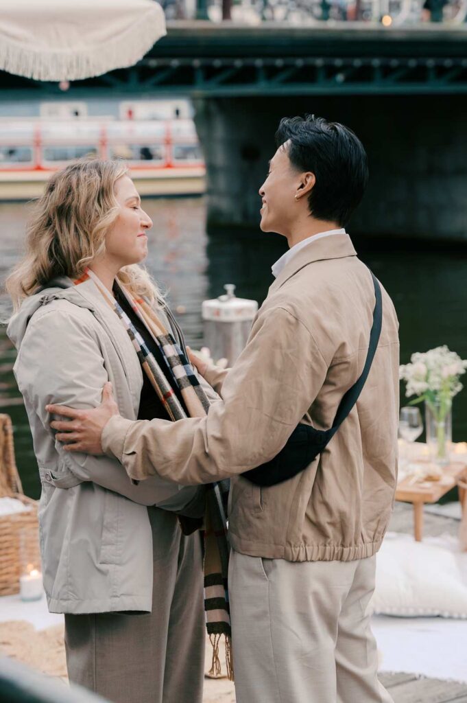 Two people stand close, smiling at each other by an Amsterdam canal, with a picnic setup in the background—a perfect moment for proposal photography.