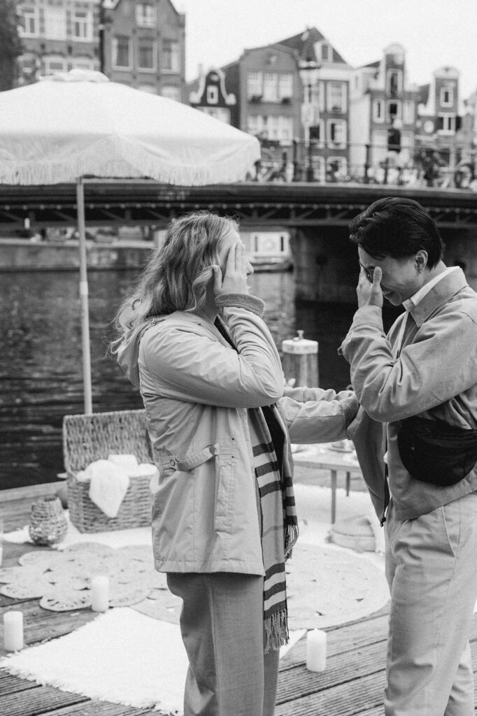 Two people stand on a dock by the Amsterdam canal, smiling and wiping away tears, sharing an emotional moment during a proposal photo shoot.