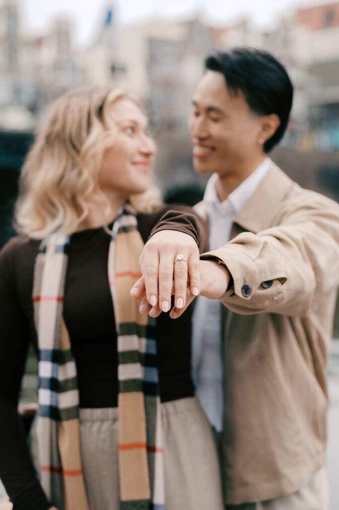 A couple smiles as the woman shows off an engagement ring on her hand, both looking happy outdoors during a surprise proposal in Amsterdam.