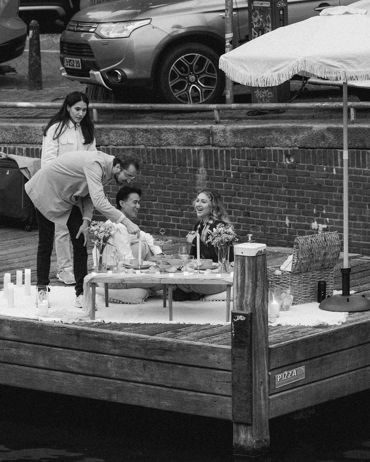 Four people enjoy a picnic on a dock by an Amsterdam canal, surrounded by flowers and a large umbrella—an idyllic setting perfect for a surprise proposal.