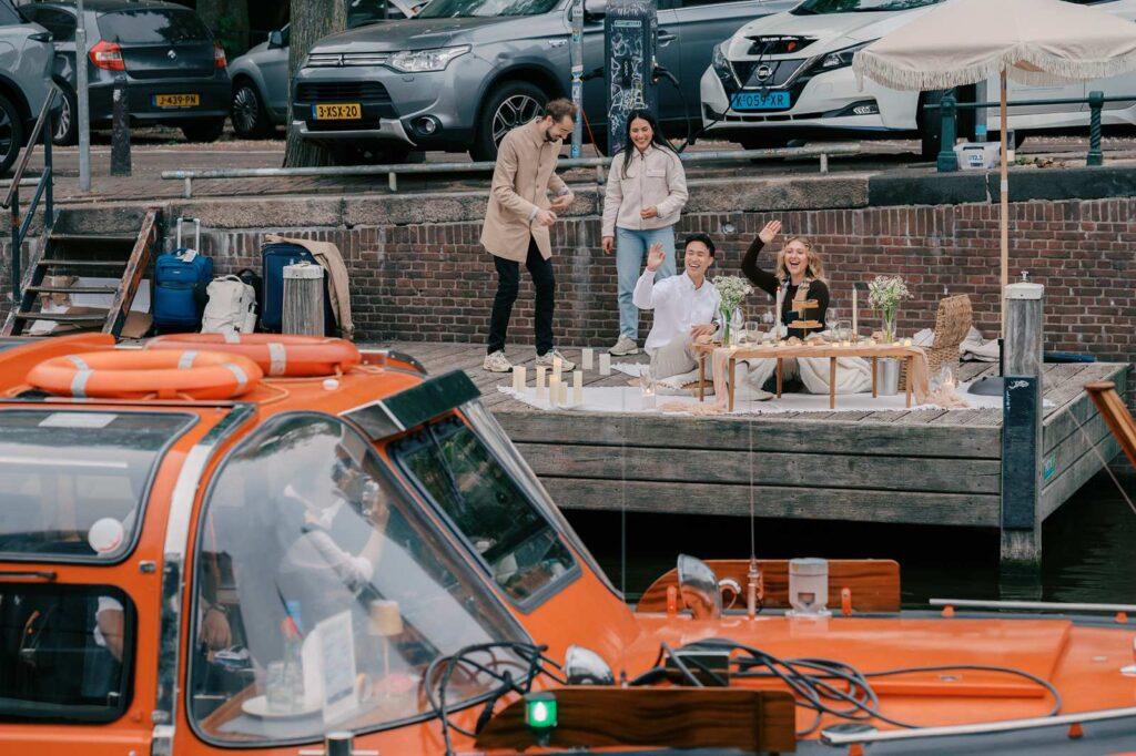Four people enjoy a picnic by an Amsterdam canal, waving at an orange boat passing in the foreground—an ideal setting for a surprise proposal or a memorable proposal photo shoot.