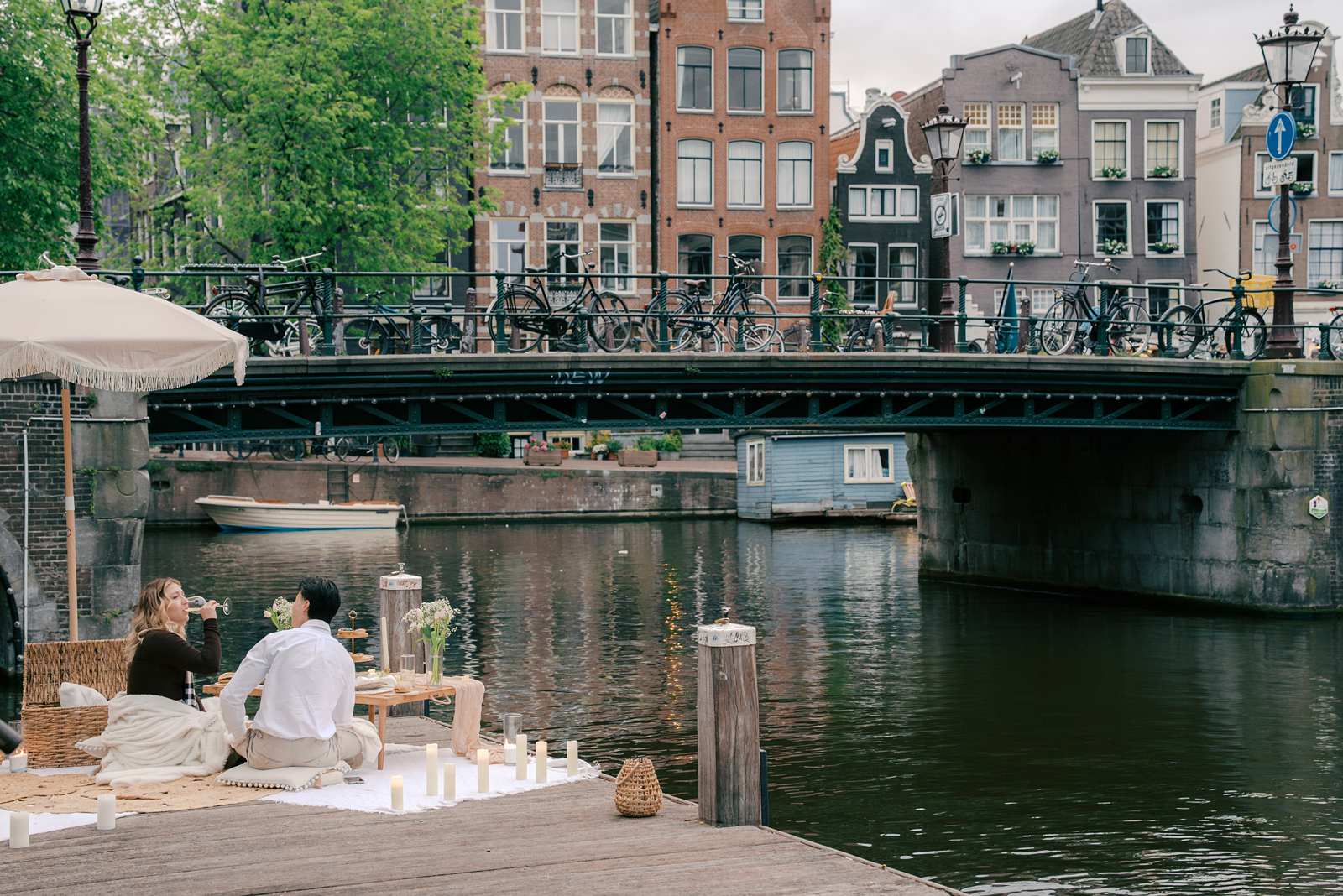 Two people enjoy a picnic by an Amsterdam canal in the city, with a bridge and bicycles in the background—perfect for candid proposal photography.