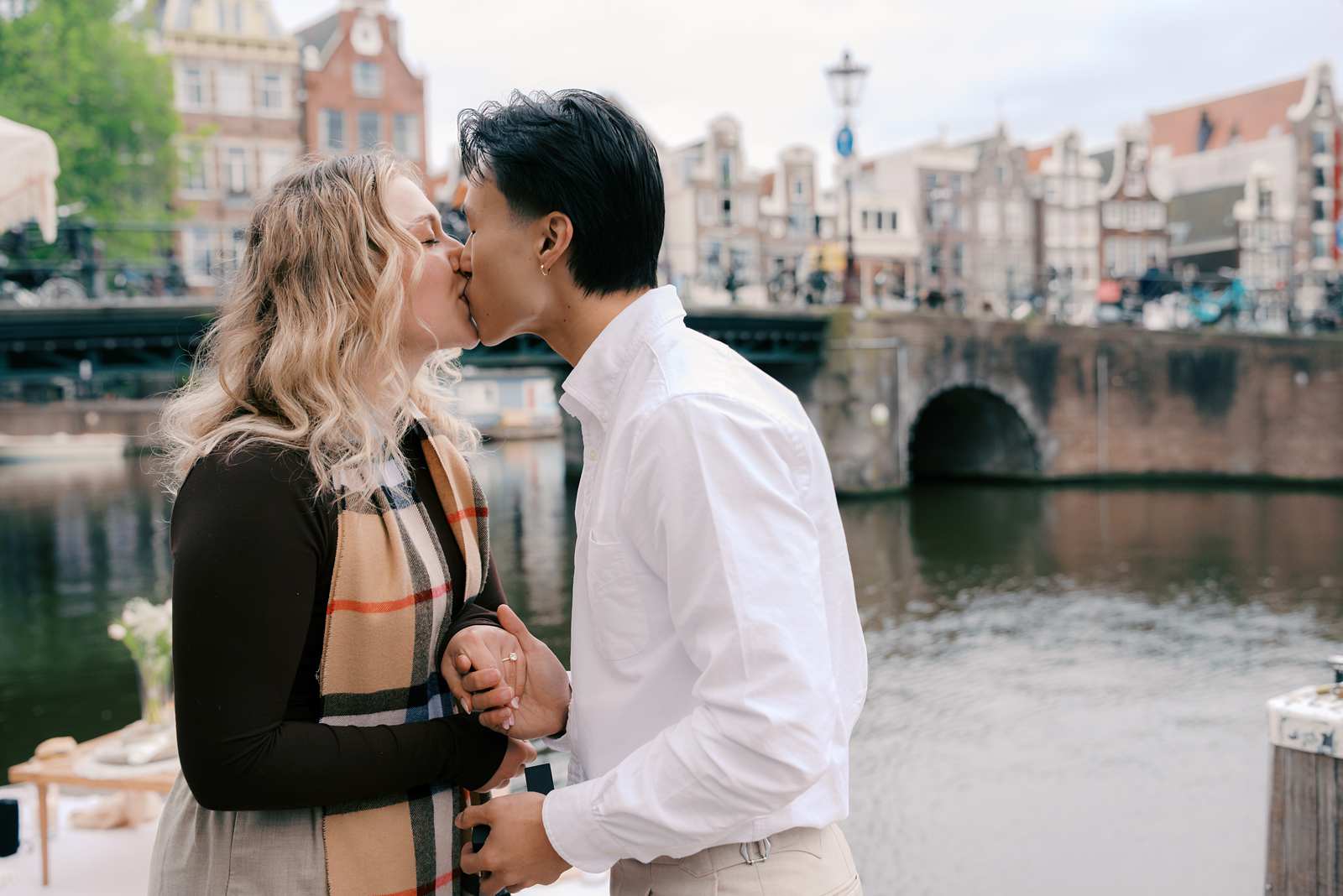 A couple kisses by a canal in Amsterdam, holding hands near a stone bridge and historic buildings, sharing a surprise proposal during a luxury picnic.
