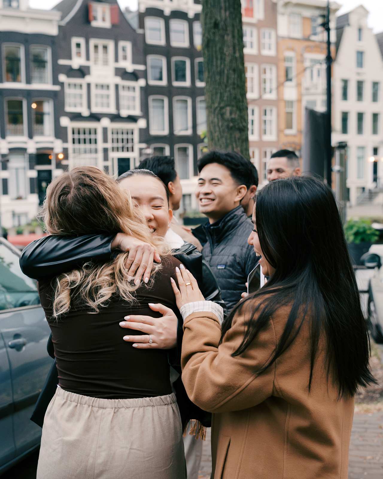 A group of friends happily hug and greet each other on a city street in Amsterdam, with tall buildings in the background.