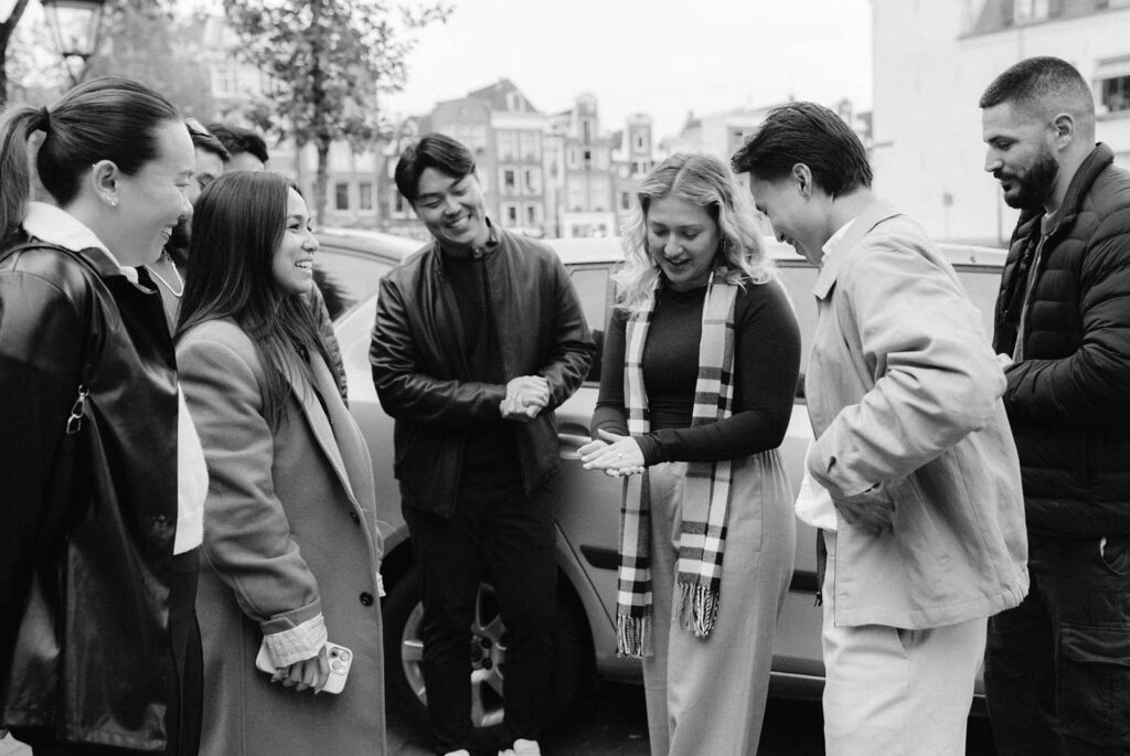 A group of people smiling and watching a woman perform a trick with her hands outdoors during a surprise proposal in Amsterdam.