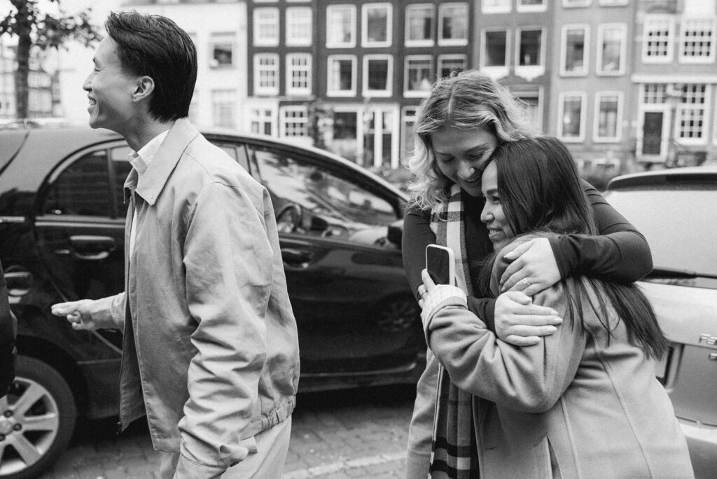 Three friends smile and hug on a city street near an Amsterdam canal, with parked cars and buildings in the background. Black and white photo.