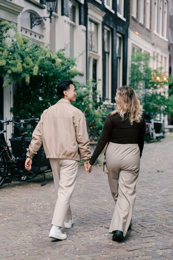 Two people hold hands while walking down a cobblestone street lined with plants and classic buildings, capturing a sweet moment that feels perfect for proposal photography in Amsterdam.