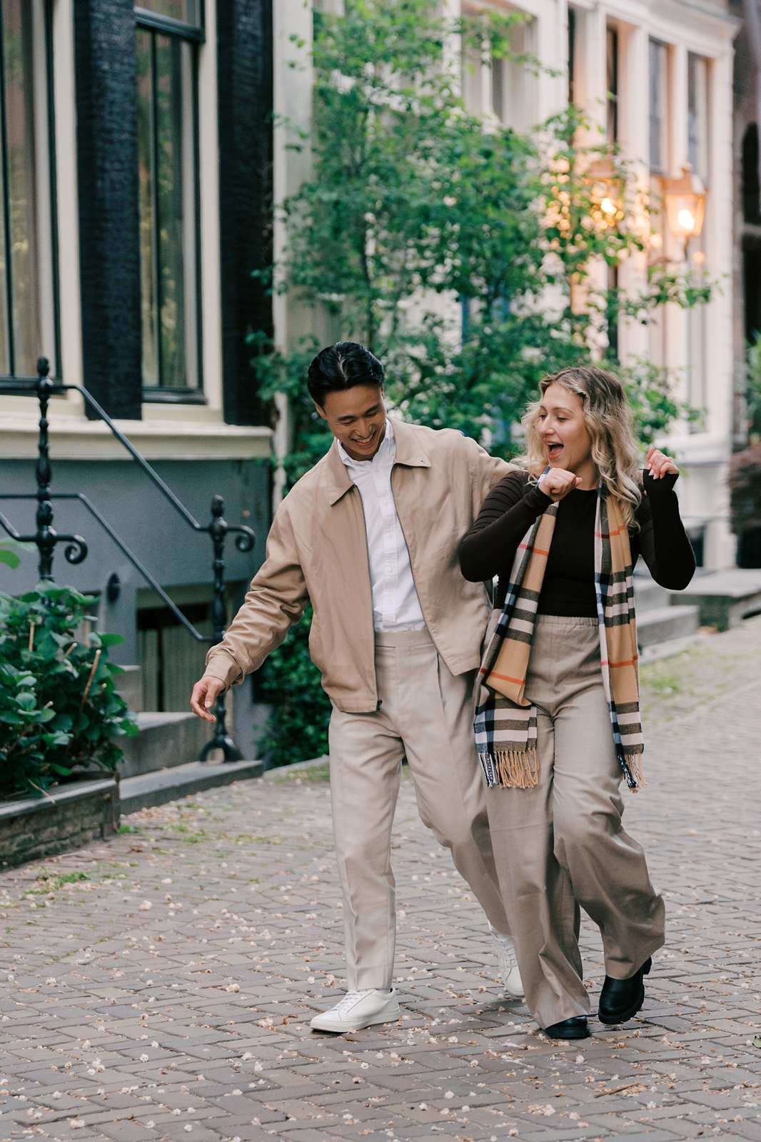 A man and woman laugh and walk together on a cobblestone street lined with plants and buildings, enjoying the charming atmosphere of Amsterdam near a scenic canal.