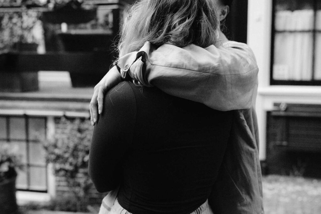 Two people embracing closely outdoors by an Amsterdam canal, one person’s arm wrapped around the other's shoulders—a tender moment perfect for proposal photography.