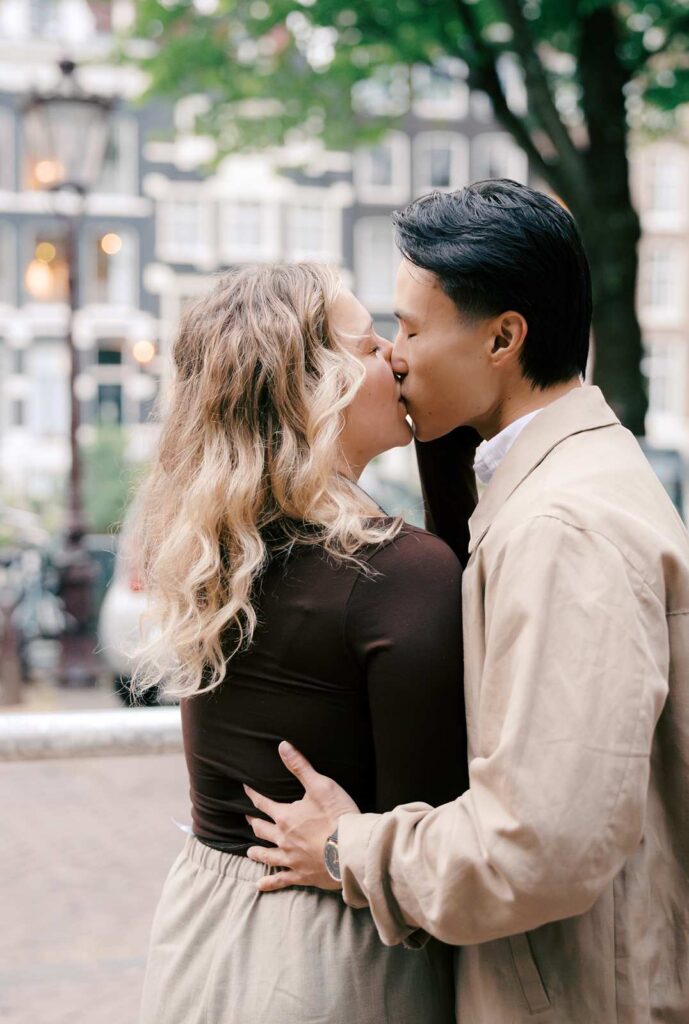 A couple shares a kiss outdoors with trees and buildings in the background, capturing a romantic moment from their proposal photo shoot.