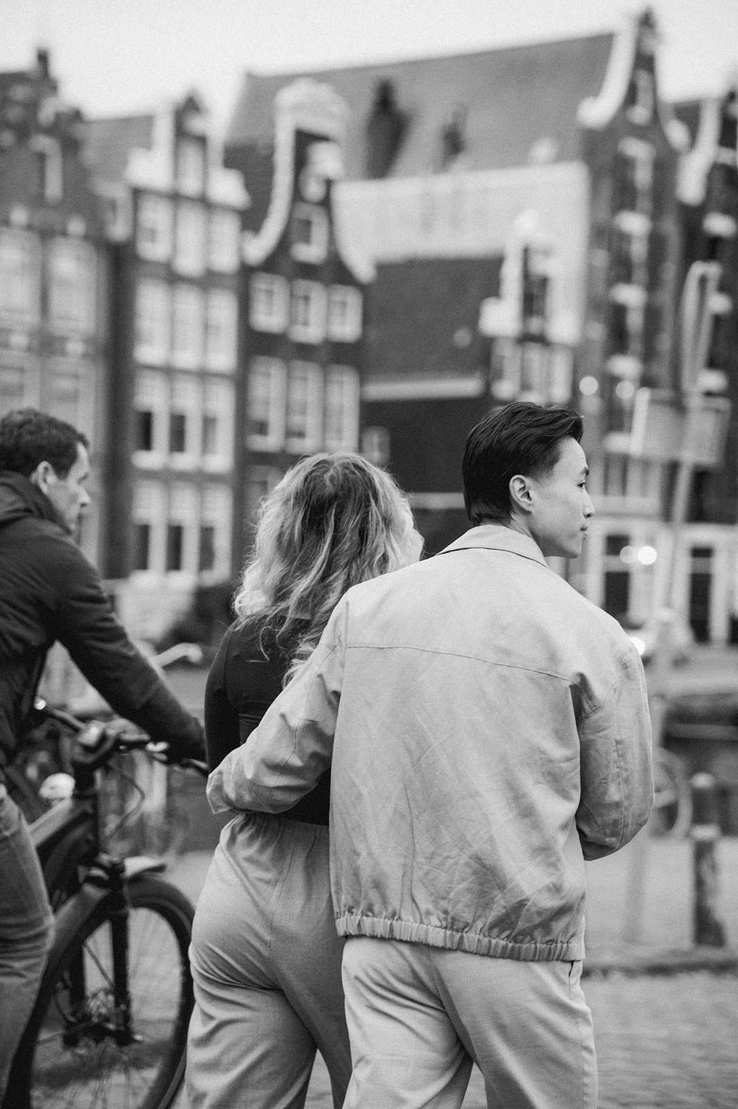 A couple walks arm-in-arm by a canal in Amsterdam while a cyclist passes by, tall buildings in the background—an ideal moment for proposal photography.