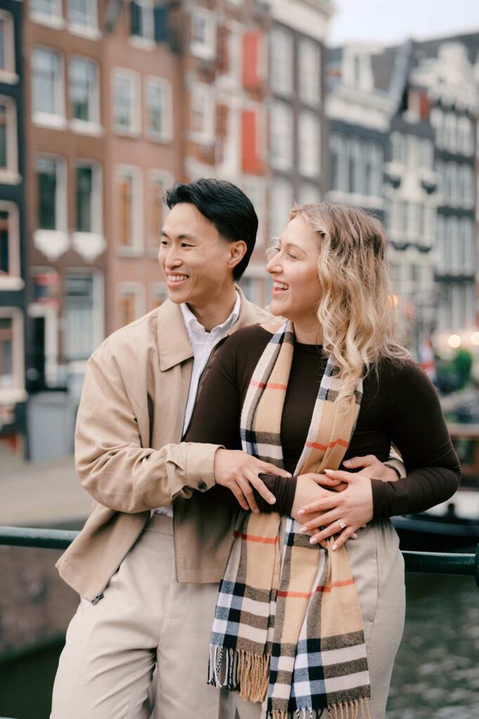 A smiling couple embraces on a city street, with charming Amsterdam canal houses in the background—capturing the joy of their surprise proposal.