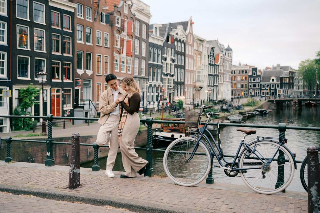 Couple embracing on an Amsterdam canal bridge with a bicycle and colorful houses in the background, perfect for a romantic proposal photo shoot.