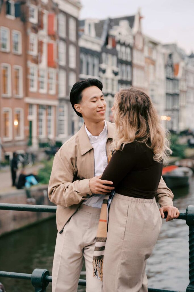 A couple stands close together on a bridge by a canal in Amsterdam, smiling at each other, surrounded by colorful buildings—the perfect moment for a proposal photo shoot.