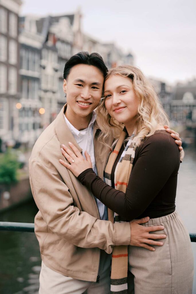 Smiling couple embracing on a bridge over an Amsterdam canal, historic buildings in the background—perfect for a proposal photo shoot.
