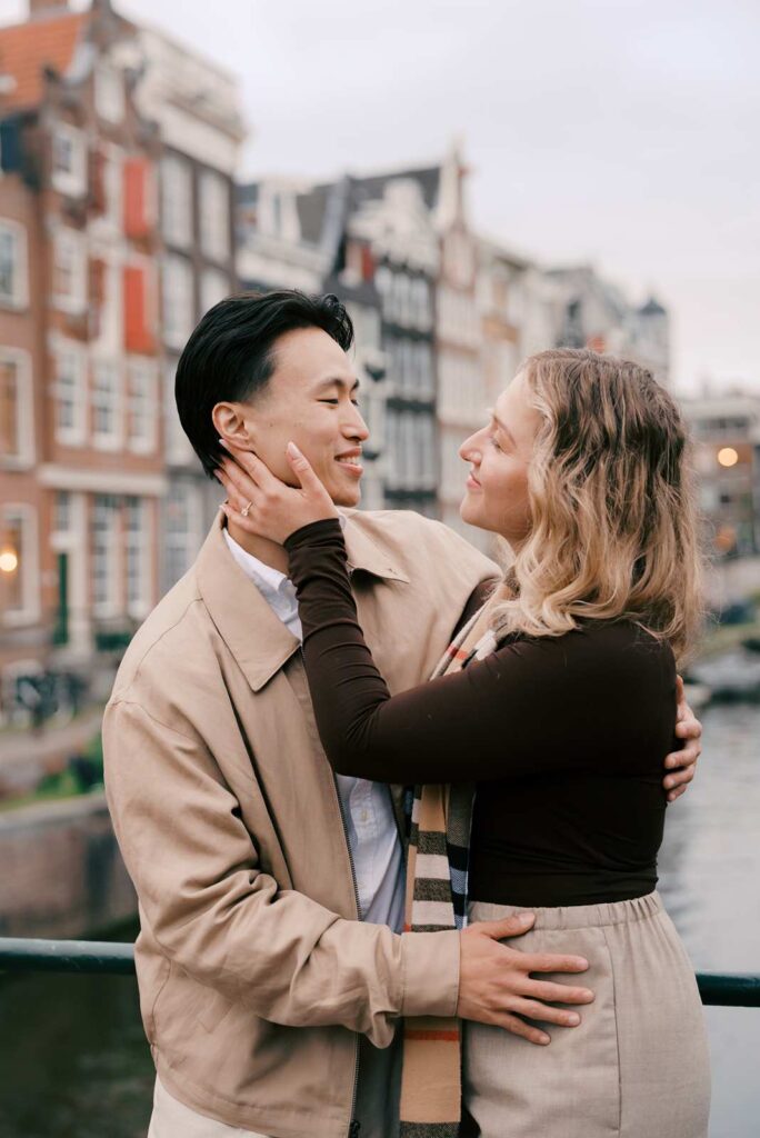 A couple embraces and smiles at each other on a bridge in Amsterdam, with canal houses in the background, capturing the joy of a surprise proposal.