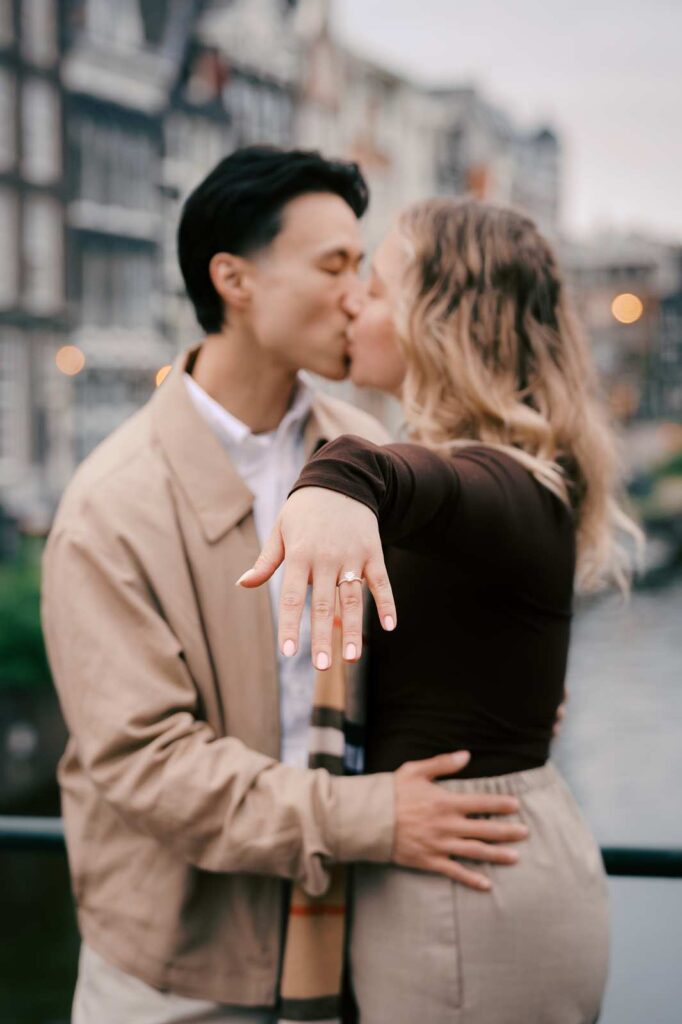 A couple kisses by an Amsterdam canal as the woman shows off her engagement ring, capturing a romantic proposal photo shoot.