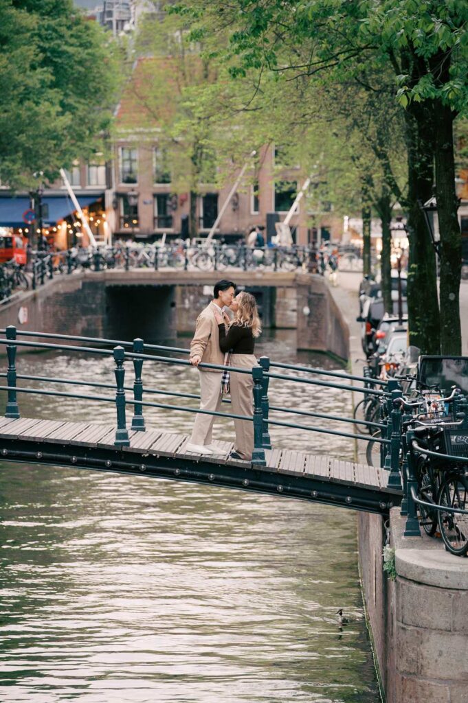 A couple kisses on a bridge over a canal in Amsterdam, surrounded by trees, bicycles, and the promise of a luxury picnic.