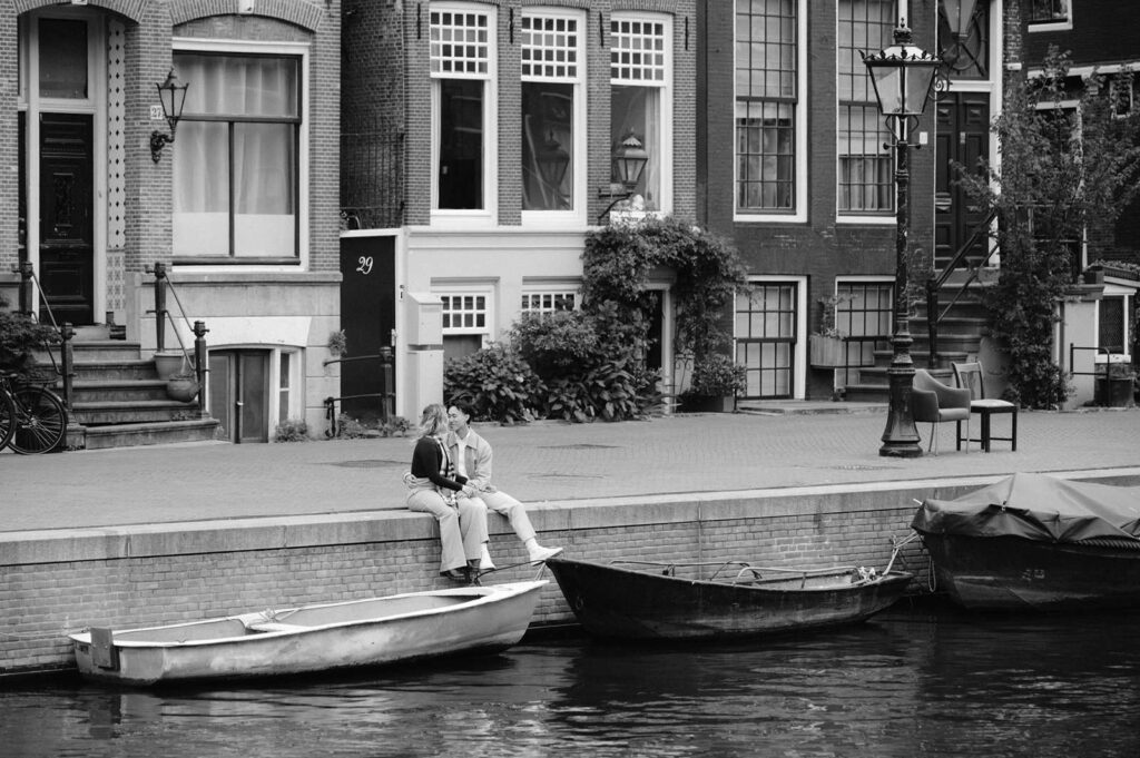 A couple sits on a canal edge in Amsterdam, sharing a kiss near boats and classic buildings—a perfect moment for proposal photography or a luxury picnic by the water.