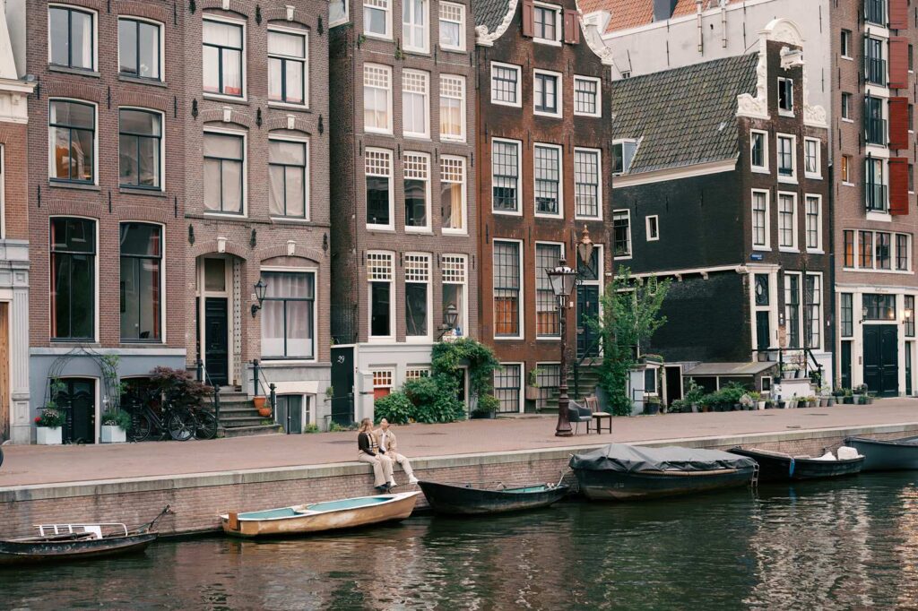 Two people sit by an Amsterdam canal in front of tall, old buildings and boats, creating a perfect scene for proposal photography.