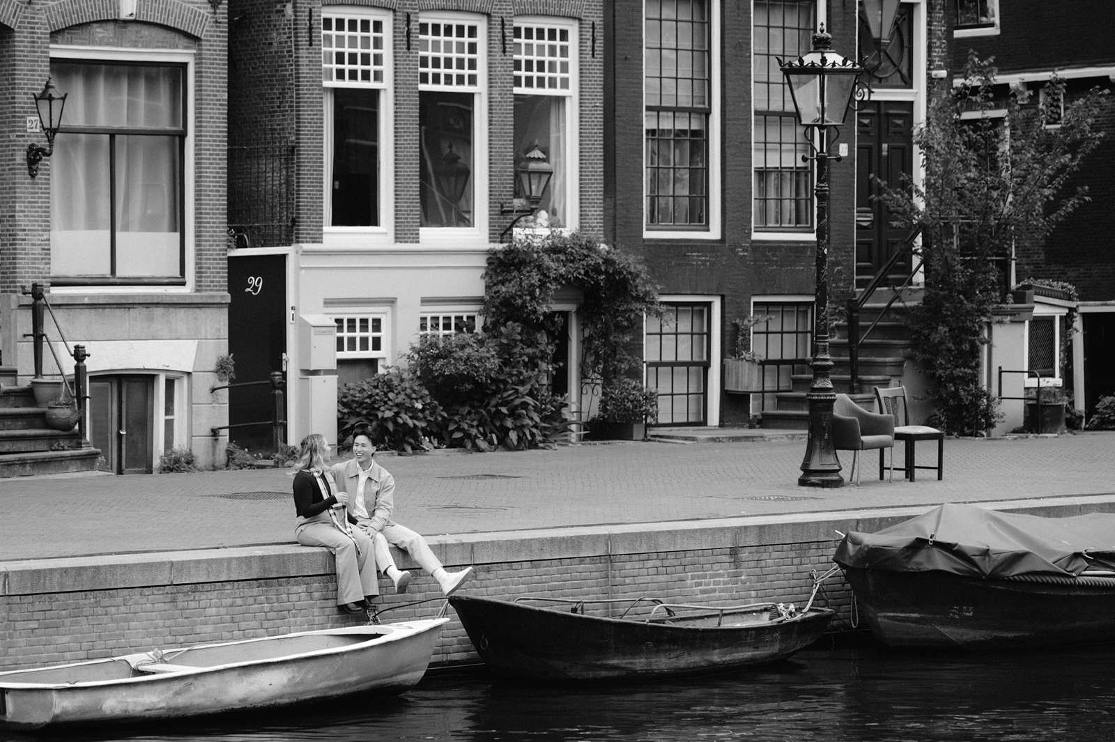 Two people sit and talk by a canal with boats, in front of traditional Amsterdam buildings in black and white—perfect for a surprise proposal or intimate proposal photo shoot.