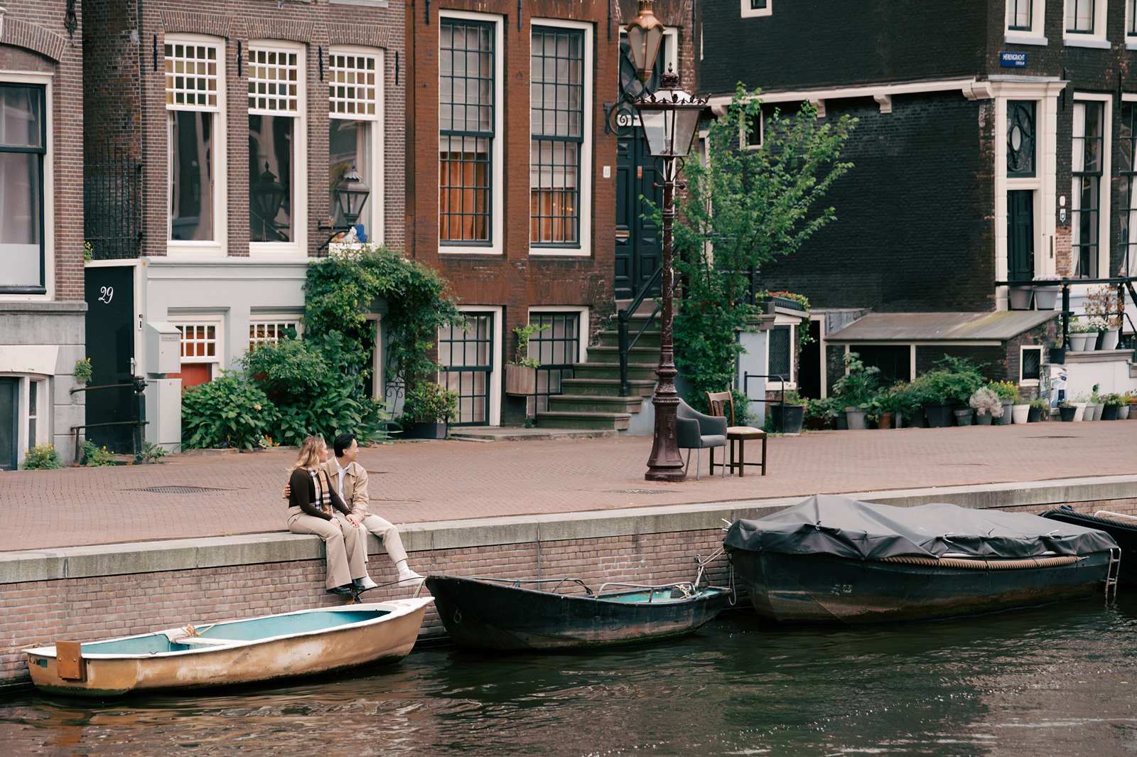Two people enjoy a luxury picnic by an Amsterdam canal in front of classic Dutch houses, with boats moored in the water nearby.
