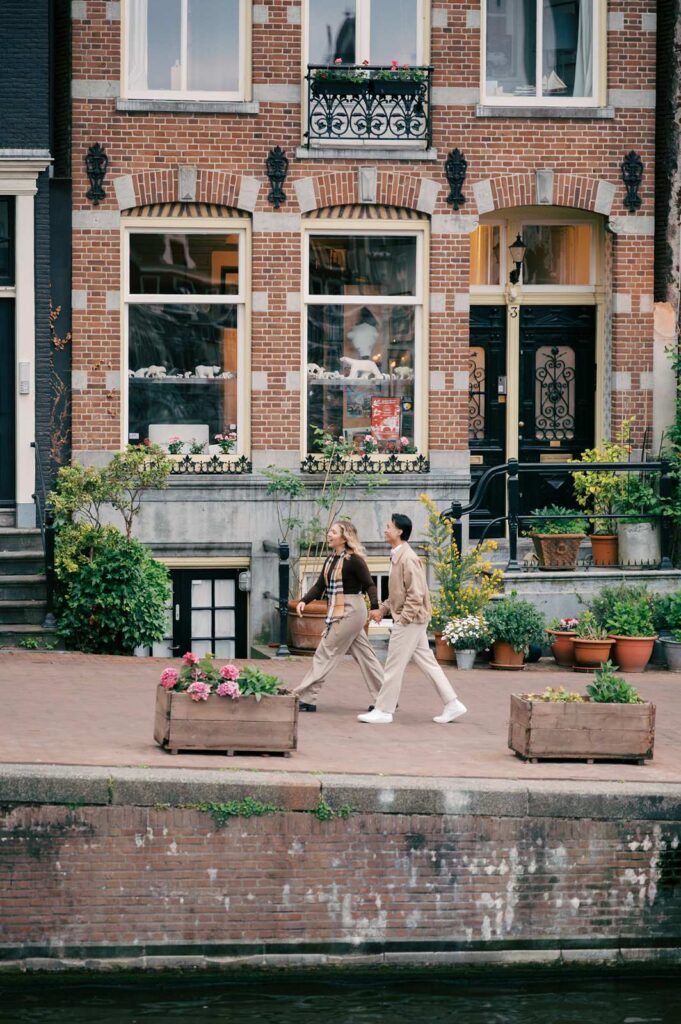 A couple walks past a brick building with potted plants along an Amsterdam canal, the perfect spot for a surprise proposal or candid proposal photography.