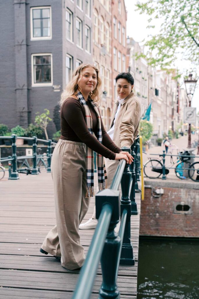 Two people stand on a bridge over a canal in Amsterdam, smiling and posing for the camera, capturing a joyful moment that could be part of memorable proposal photography.