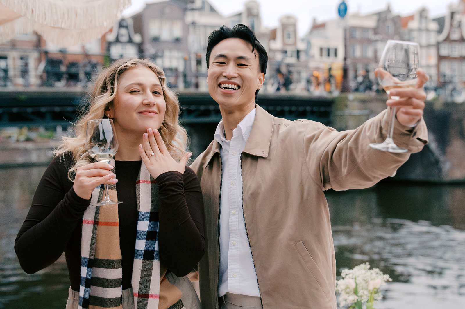 Smiling couple holding wine glasses by an Amsterdam canal, with buildings and a bridge in the background—a perfect moment for proposal photography.