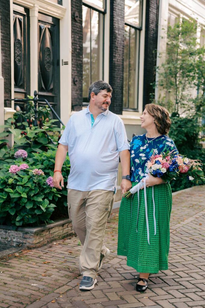 A smiling couple holding hands walks together on a cobblestone street lined with plants, capturing memories during their anniversary photo session.