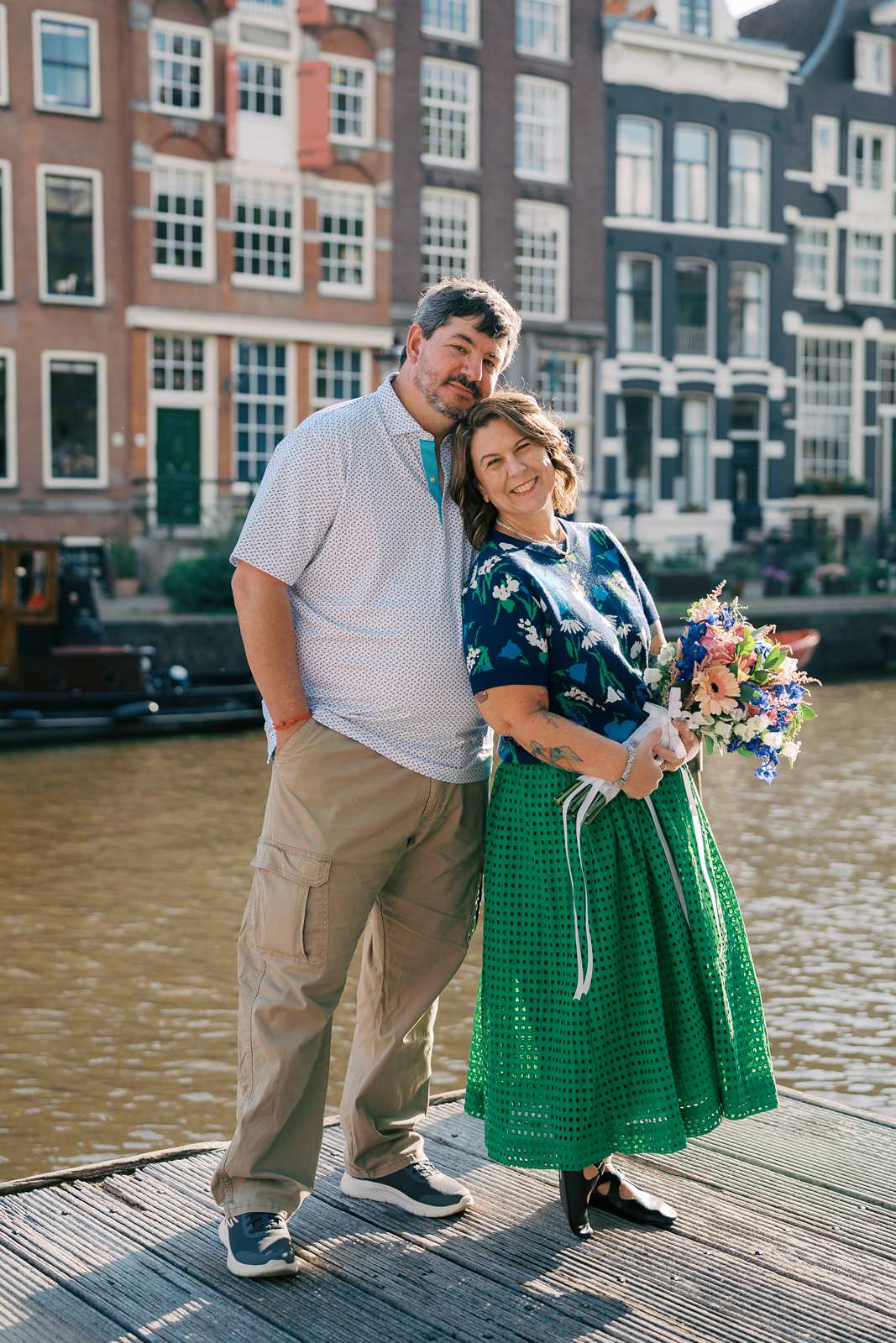 A smiling couple stands by a canal during their Amsterdam photo shoot; a woman holds a bouquet and leans against her husband.
