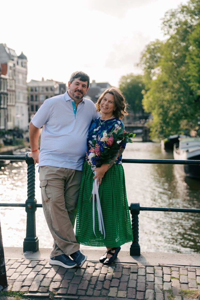 A couple poses and smiles by a canal with trees and buildings in the background, celebrating their anniversary in Amsterdam with a golden hour photo shoot
