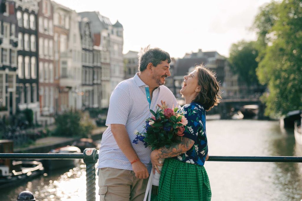 A joyful couple on a bridge during their Amsterdam photo shoot, holding flowers and laughing together
