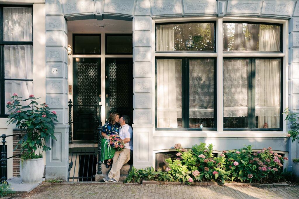 A couple stands in a doorway in Amsterdam, holding a custom bouquet and kissing during their intimate anniversary photo session to celebrate in Amsterdam.